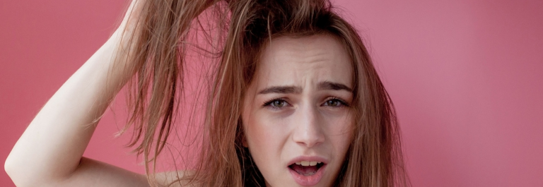 woman with disgruntled face holding waxy & greasy hair after shower against pink background