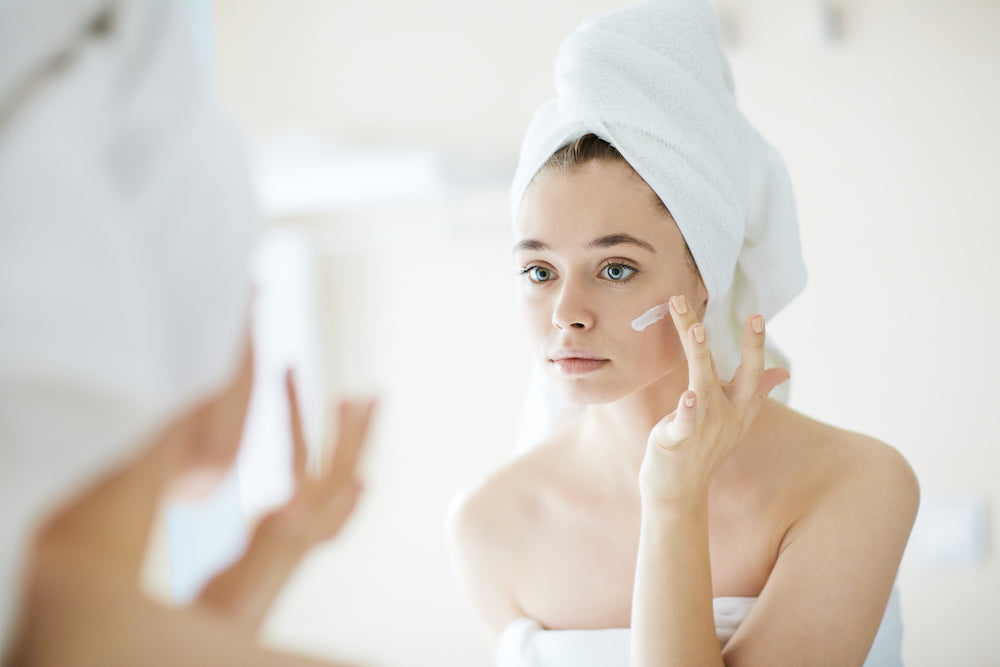 Woman wearing a towel on her head and applying cream to her face.